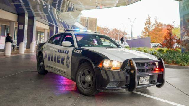 A black and white Dallas police car with lights on sits under the overhang of a large building.
